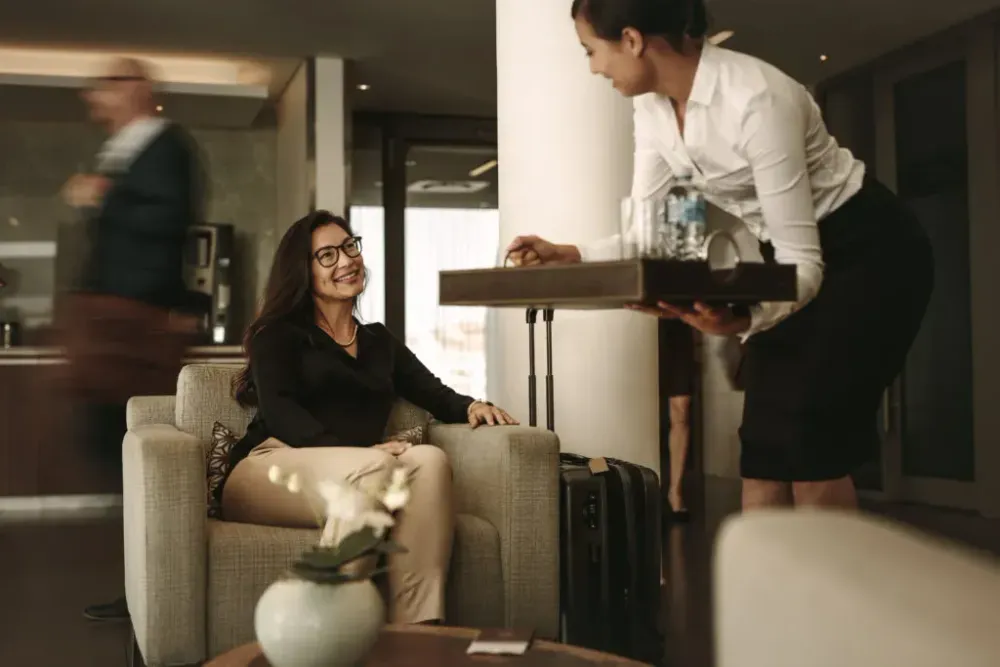 Woman sitting next to her luggage in an airport lounge while another woman offers her drinks from a tray